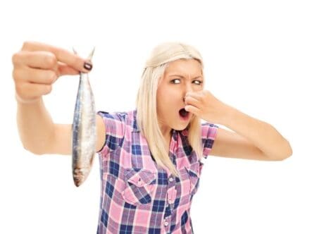 A girl is holding a fish in her hand and covering her nose with her other hand due to the smell of the fish.